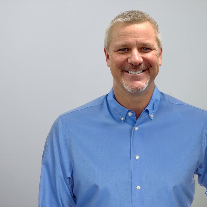 B. Woerner_withTorso_Light Background Bert Woerner wearing a blue button up shirt in front of a grey background.